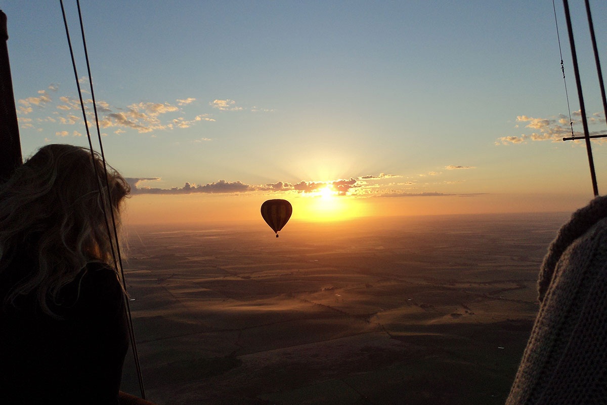 Person in hot air balloon above clouds, looking out towards another hot air balloon; sunset in background