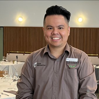 Ben Tan faces the camera smiling. He has brown hair and brown eyes and wears a formal grey button shirt. In the background is a restaurant. There's tables with white table cloths draped over the top. Glassware is placed on the tables fashionably. There are chairs tucked into the tables neatly. On the back wall, the bottom half if covered with wood whilst the top half is a white wall.