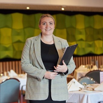 Student Camille facing the camera holding a clipboard. She stands in an empty restaurant with the tables set for service, with a white tablecloth and cutlery. Behind her is a green wall feature and wooden pannels.