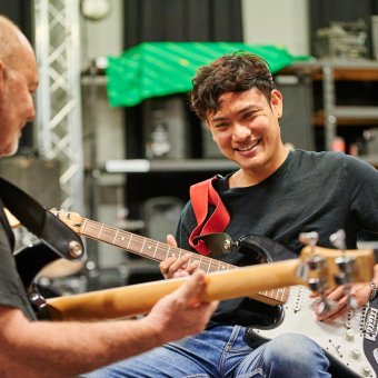 Student Farook (George) is seated holding an electric guitar. He faces a man who also holds an electric guitar. Farook is smiling at the instructor as he strums.