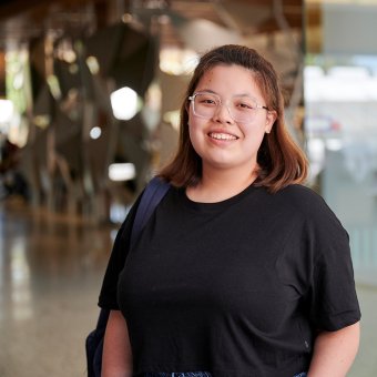 Student Athena standing in foyer of Perth TAFE campus. She wears a black t-shirt and a backpack on one shoulder. She faces towards the camera with a big smile.