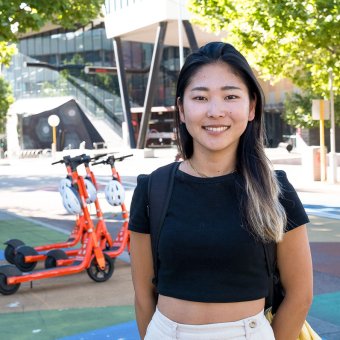 Student Zaki at North Metropolitan TAFE. She stands, facing the camera. In the background are several orange e-scooters and the NMTAFE campus. There are green trees to the right of her in the background.