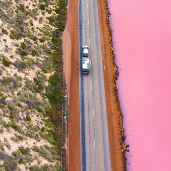 Car drives on road between green hills and a pink lake