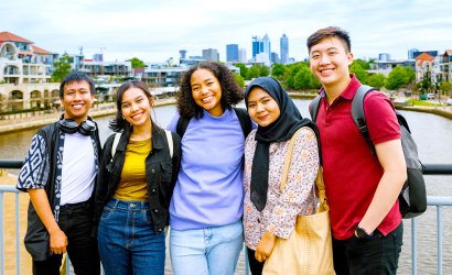 Five students stand together, facing the camera with big smiles. In the background is a body of water and a scenic view of the Perth skyline
