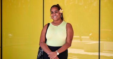 Student Keloha stands facing the camera with a big smile. She wears a yellow top with a yellow frangipani tucked behind her ear. Behind her is a yellow wall.
