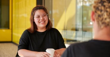 Student sitting at table with a coffee in her hands. She smiles, facing towards another student who is out of focus. Behind the student is a yellow wall and window.