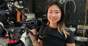 Student Zaki, standing behind a professional camera secured on a tripod. She holds the tripod handle with her right hand. Zaki faces the camera, with a big smile. She's inside, with cords and equipment in the foreground.