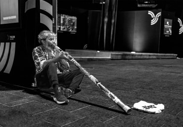 Indigenous Australian man sits on the floor playing a didgeridoo. The image is in black and white.