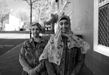 Two females face towards the camera. The woman closest to the camera is wearing a hijab. Both women are smiling. In the background there's trees, buildings and a road with no cars.