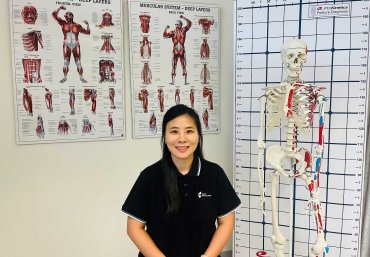 Lecturer Jinjoo Park sits in the centre of the photo wearing her South Metropolitan TAFE work polo shirt. The photo has been taken inside a room. The white wall behind her has two large posters that show diagrams of different muscles on the human body. On the right of the image is a skeleton.