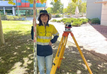 Student Sachiho stands under the shade, hand on hip and wearing a long sleeve hi vis work shirt. She's holding a piece of surveying equipment. Beside her is a tripod with a Robotic Total Station. She smiles towards the camera.